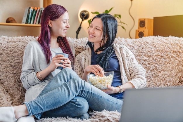 Giovani ragazze felici che bevono caffè e mangiano popcorn mentre guardano lo spettacolo sul computer portatile