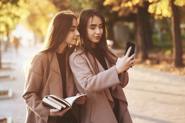 Giovani ragazze gemelle piuttosto mora che si guardano e fanno selfie con il telefono nero, mentre una di loro tiene in mano libri, indossa un cappotto, in piedi al vicolo del parco soleggiato autunnale su sfondo sfocato.