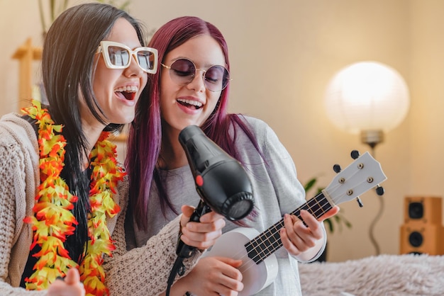 Giovani ragazze gioiose con gli occhiali da sole che ballano e cantano con l'asciugatrice e l'ukulele a casa