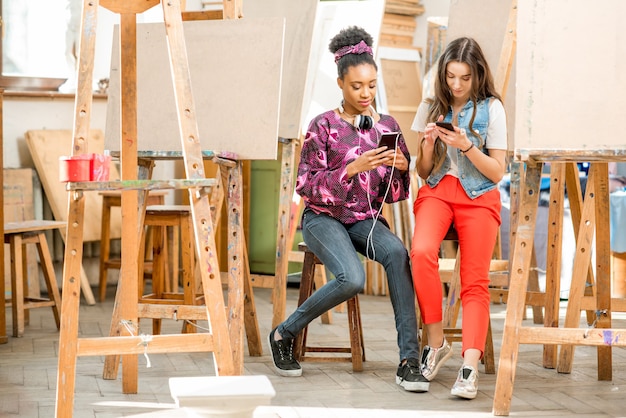 Giovani studenti creativi multietnici seduti con i telefoni durante la pausa nello studio universitario per dipingere