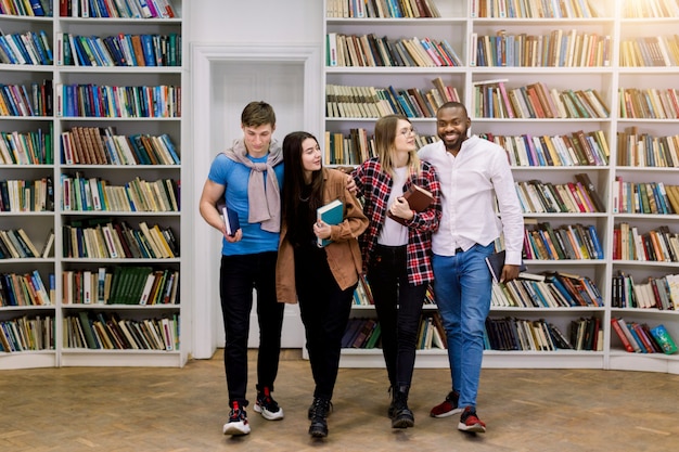 Giovani studenti multietnici, ragazze e ragazzi in possesso di libri, abbracciati, in piedi in biblioteca sullo spazio degli scaffali dei libri