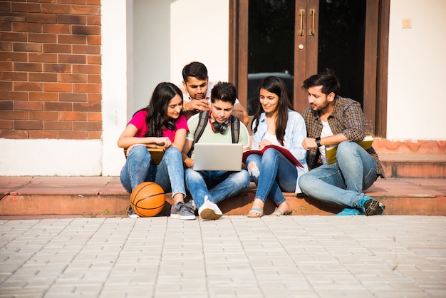 Giovani studenti universitari indiani asiatici che leggono libri, studiano sul laptop, si preparano per l'esame o lavorano su un progetto di gruppo mentre sono seduti sull'erba, sulle scale o sui gradini del campus universitario
