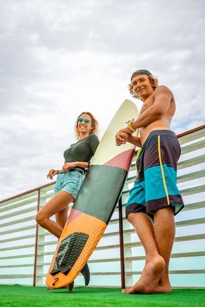 Giovani surfisti attivi sorridenti delle coppie che si rilassano sulla spiaggia dopo lo sport con il surf. Uno stile di vita sano. Sport acquatici estremi