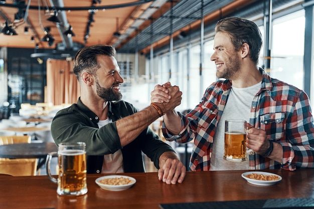 Giovani uomini allegri in abiti casual che si stringono la mano e bevono birra mentre trascorrono del tempo al pub