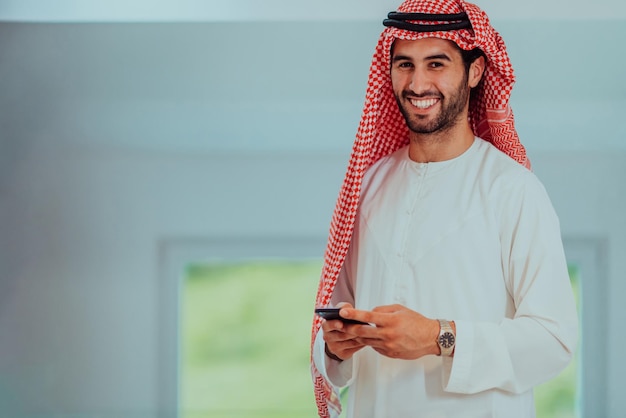 Giovani uomini d'affari arabi musulmani moderni che indossano abiti tradizionali mentre usano gli smartphone a casa.