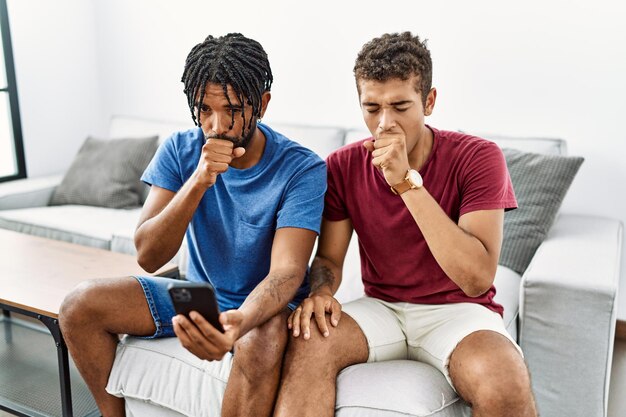 Giovani uomini ispanici che usano lo smartphone seduti sul divano di casa sentendosi male e tossendo come sintomo di raffreddore o bronchite. concetto di assistenza sanitaria.