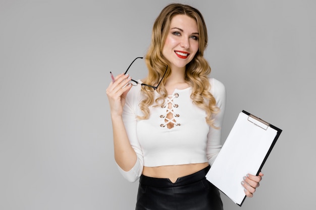 Giovani vestiti biondi attraenti della donna di affari in bianco e nero che sorridono mostrando lavagna per appunti in sue mani che stanno nel gray dell'ufficio