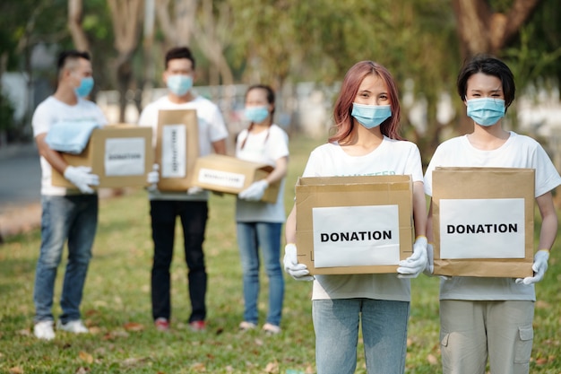 Giovani volontari asiatici positivi in maschere mediche che tengono scatole di cartone di cibo e vestiti per le persone che hanno perso il lavoro a causa della pandemia di coronavirus