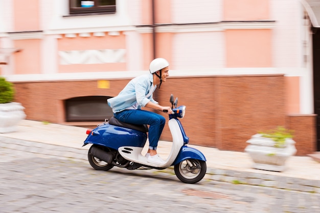 Giro in scooter. Vista laterale del giovane in casco in sella a uno scooter lungo la strada e sorridente