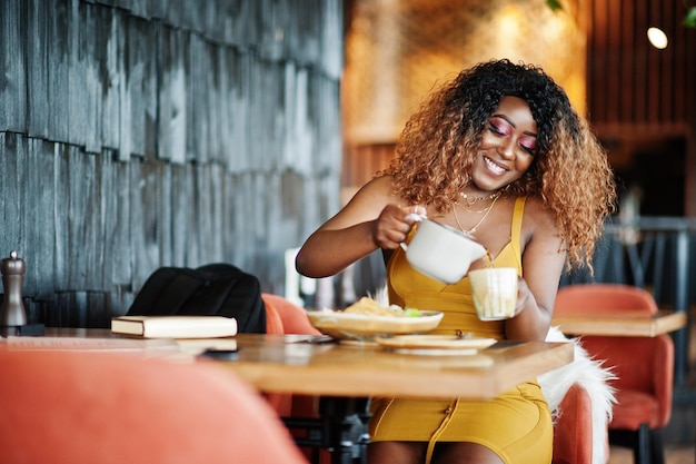 Glamour donna afroamericana in abito giallo seduto al ristorante e versa il tè dalla teiera