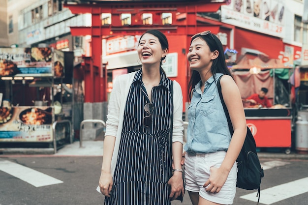 Gli amici delle ragazze asiatiche viaggiano in Giappone si trovano su una strada urbana di fronte al grande cancello rosso di torii. due giovani donne allegre sorridenti visitano un famoso sito storico. carrello del venditore ambulante in background sul mercato cittadino.