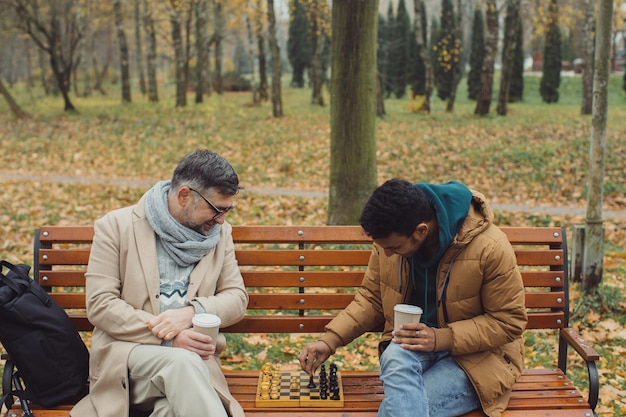 Gli amici giocano a scacchi su una panchina in un parco autunnale Amicizia multiculturale di persone di età diverse