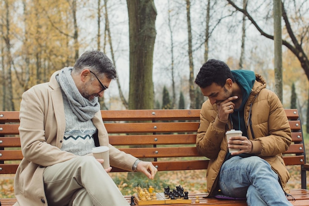Gli amici giocano a scacchi su una panchina in un parco autunnale Amicizia multiculturale di persone di età diverse