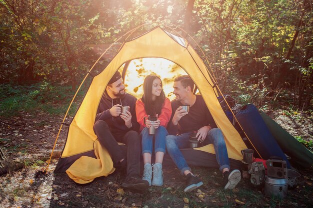 Gli amici si siedono in tenda e si parlano. Tengono tazze termiche con bevanda calda. Sguardo della giovane donna al ragazzo a destra. Lei sorride.