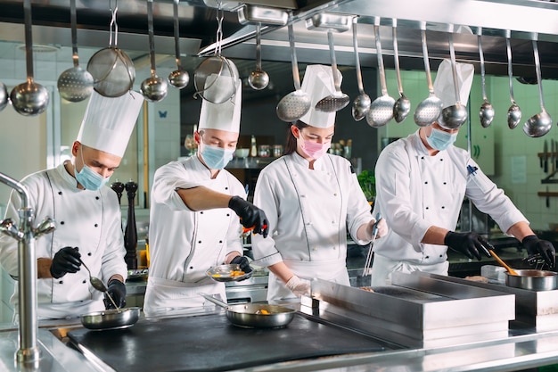 Gli chef con maschere e guanti protettivi preparano il cibo nella cucina di un ristorante o hotel.