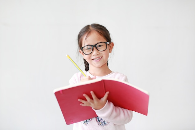 Gli occhiali da portare della bambina felice e tengono un libro e scrivono con la matita su bianco.