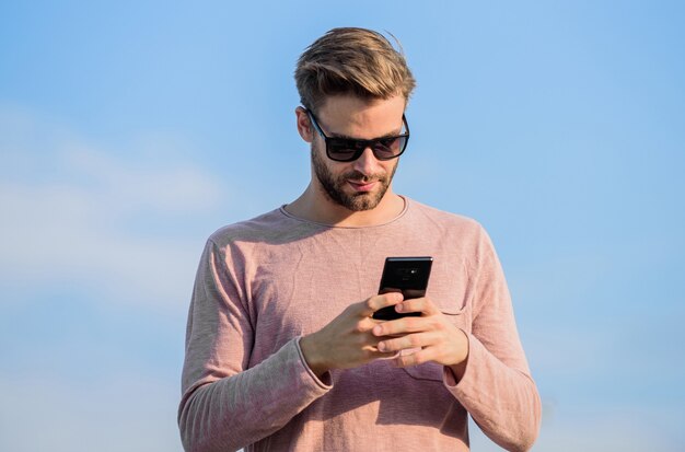 Gli occhiali da sole di usura dei pantaloni a vita bassa tengono il fondo del cielo del telefono cellulare. Chiamata da smartphone hipster. Chiamata mobile uomo. Rimani in contatto. Concetto di chiamata mobile. Importante conversazione mobile. Applicazione e Internet.