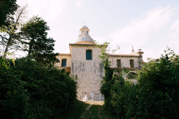 Gli sposi camminano mano nella mano accanto a una bellissima vecchia chiesa tra gli alberi