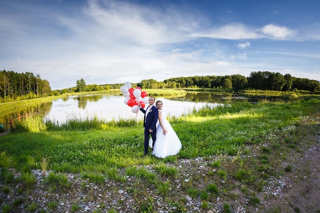 Gli sposi nella natura vicino al lago con palloncini bianchi rossi La sposa abbraccia lo sposo Panorama del matrimonio Il giorno più bello di una giovane coppia