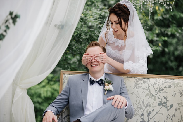 Gli sposi sono seduti su un bellissimo divano in un gazebo in giardino. Tenda bianca. Fogliame verde. La sposa in un vestito soffice rosa, lo sposo in un vestito grigio.