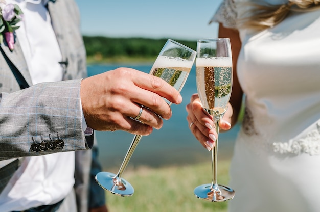 Gli sposi tengono in mano un bicchiere di champagne e stanno sulla natura durante la cerimonia nuziale. Avvicinamento. Vacanza. Guarda gli occhiali. Pane abbrustolito.