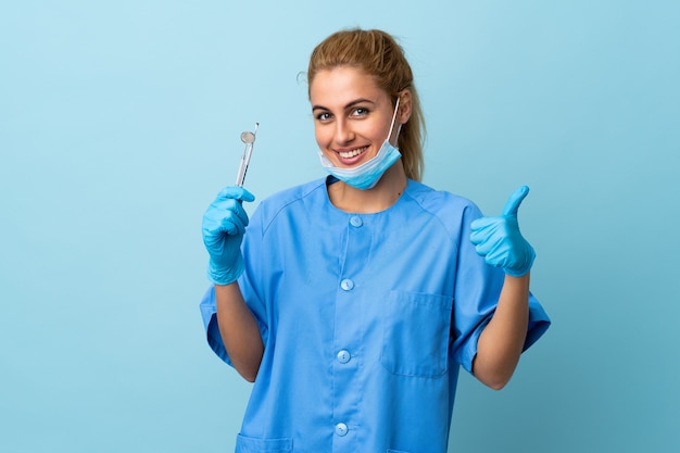 Gli strumenti di tenuta del dentista della giovane donna sopra il blu isolato dare pollici aumentano il gesto
