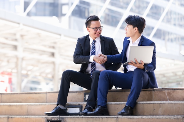 Gli uomini d&#39;affari asiatici due che si siedono stanno lavorando al computer portatile in città