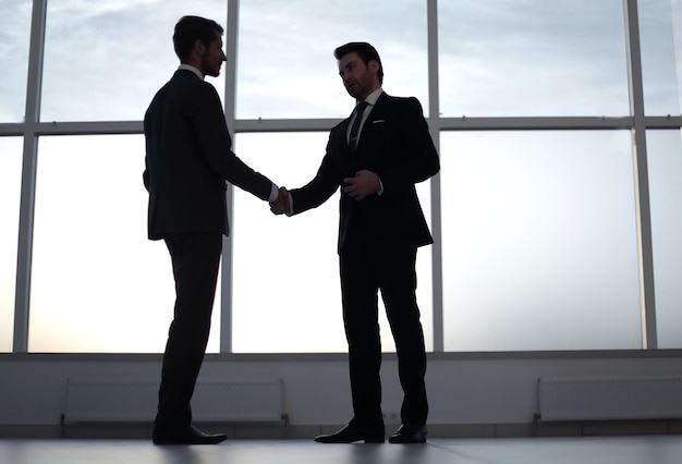 Gli uomini d'affari si stringono la mano in piedi vicino a una grande finestra dell'ufficio il concetto di cooperazione