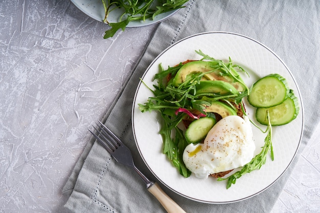 Gli uova affogate con le erbe, l'avocado ed il cetriolo sono servito su un piatto su un fondo leggero