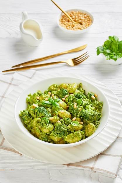 Gnocchi italiani con cimette di broccoli conditi con pesto di basilico e salsa di formaggio cremoso cosparso di pinoli in una ciotola bianca su un tavolo di legno, primo piano, vista verticale dall'alto