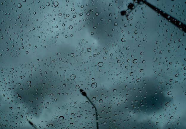 Gocce di pioggia su vetro trasparente contro sfocatura cielo tempestoso scuro e palo elettrico.