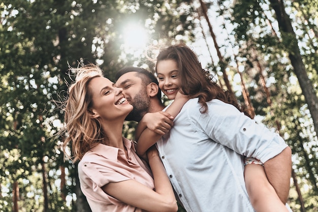 Godendo del tempo insieme. Giovane padre amorevole che porta sua figlia sorridente e bacia sua moglie mentre trascorre il tempo libero all'aperto