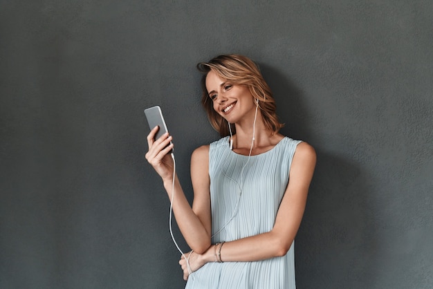 Godendo della musica preferita. Attraente giovane donna in possesso di uno smartphone e sorridente in piedi su sfondo grigio gray