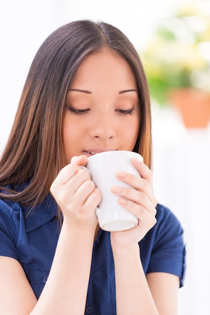 Godendo di bevanda calda. Bella giovane donna asiatica che beve una bevanda calda mentre è seduta al chiuso