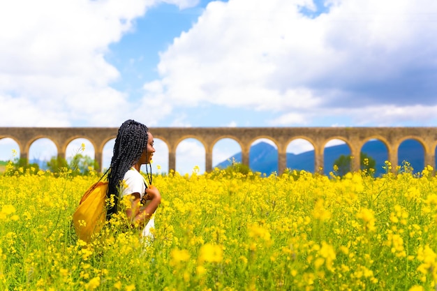 Godendo di una ragazza etnica nera primaverile con un viaggiatore di trecce in un campo di fiori gialli