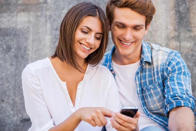 Godendo il loro tempo libero insieme. Bella giovane coppia di innamorati seduti vicini l'uno all'altro mentre la donna punta il telefono cellulare e sorride