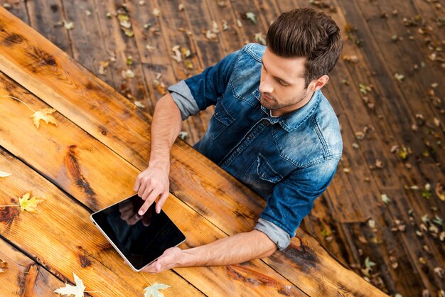 Godendo il suo lavoro all'aperto. Vista dall'alto di un giovane fiducioso che lavora su un tablet digitale mentre è seduto al tavolo di legno all'aperto