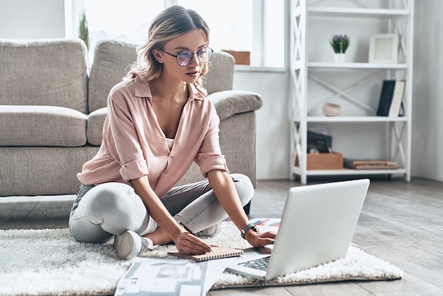 Godendo il suo lavoro creativo. Giovane donna premurosa in occhiali che lavora usando il computer mentre si pavimenta a casa