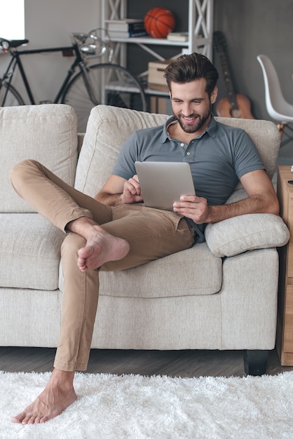 Godendo il suo tempo libero a casa. Bel giovane che usa la sua tavoletta digitale con un sorriso mentre è seduto sul divano di casa