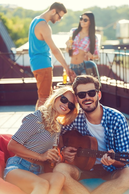 Godendo il tempo con gli amici. Bella giovane coppia che si lega l'una all'altra e si siede sul sacchetto di fagioli con la chitarra mentre due persone fanno il barbecue in sottofondo