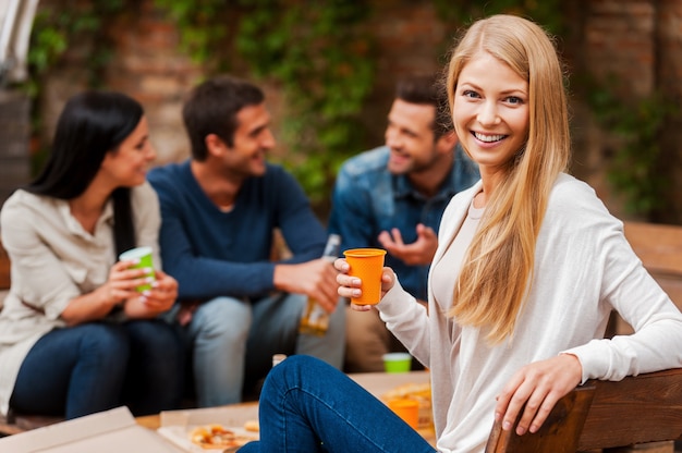 Godendo il tempo con gli amici. Sorridente giovane donna in possesso di vetro e guardando la telecamera mentre i suoi amici parlano tra loro in background