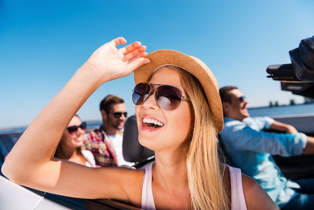 Godendo il viaggio su strada. Un gruppo di giovani felici che si godono il viaggio nella loro decappottabile mentre la bella donna si aggiusta il cappello e sorride