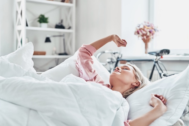 Godendo la domenica mattina. Attraente giovane donna che tiene gli occhi chiusi e si allunga mentre giace a letto a casa