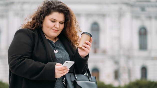 Godendo la passeggiata Donna taglie forti Comunicazione online Stile di vita virtuale Signora grassa allegra e contenta che tiene il caffè da asporto digitando il telefono cellulare sulla vista della strada