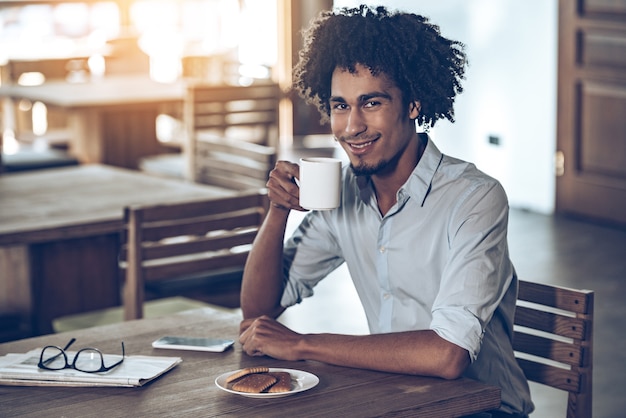 Godendo la sua mattinata. Giovane africano che tiene tazza di caffè e guarda la telecamera con un sorriso