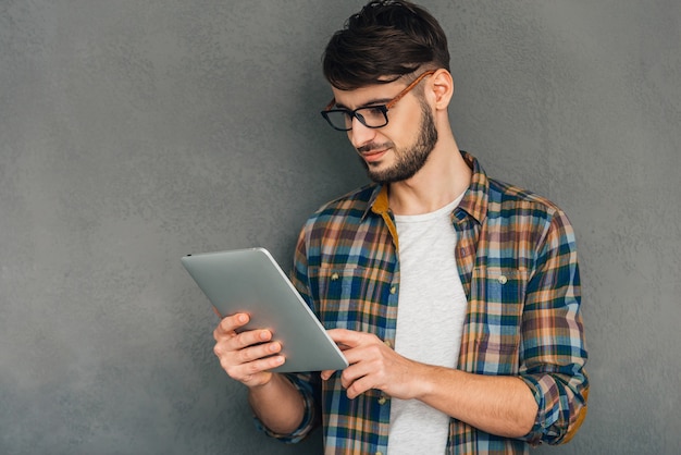 Godendo la sua tavoletta digitale. Fiducioso giovane che tiene la sua tavoletta digitale e la guarda mentre si trova in piedi su uno sfondo grigio