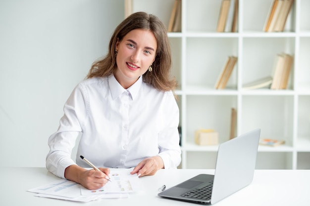 Godersi il lavoro Donna felice Routine quotidiana Elegante signora sorridente in camicia bianca seduta scrivania aperto laptop che controlla i documenti all'interno della stanza luminosa