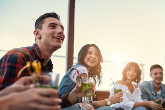 Godersi il tramonto giovani e felici che chiacchierano e bevono cocktail seduti sul tetto