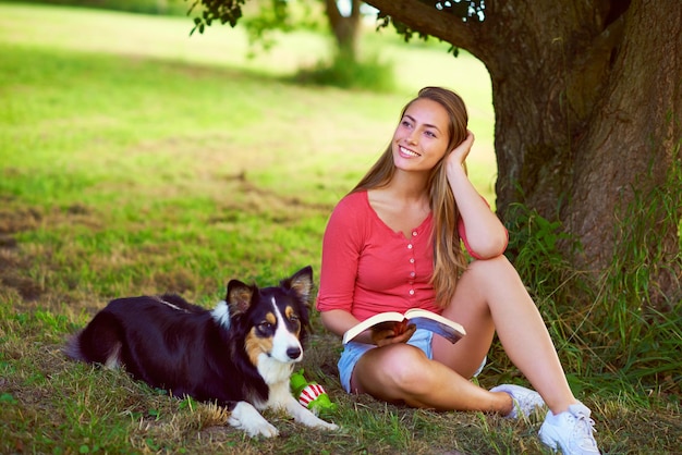 Godersi una giornata spensierata al parco Inquadratura di una giovane donna che legge un libro mentre è seduta con il suo cane sotto un albero