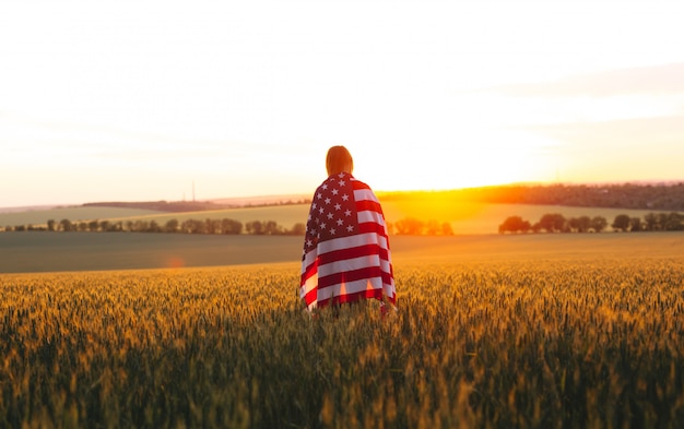 Goditi la donna con la bandiera americana in un campo di grano al tramonto. 4 luglio. Festa dell'indipendenza, festa patriottica.